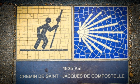 Wegweiser an einer Hauswand mit dem Piktogramm eines Pilgers und gelber Muschel für den Jakobsweg am 15. Juli 2019 in Chartres. Darunter ist die Entfernung nach Santiago de Compostela mit 1625 Km angegeben.